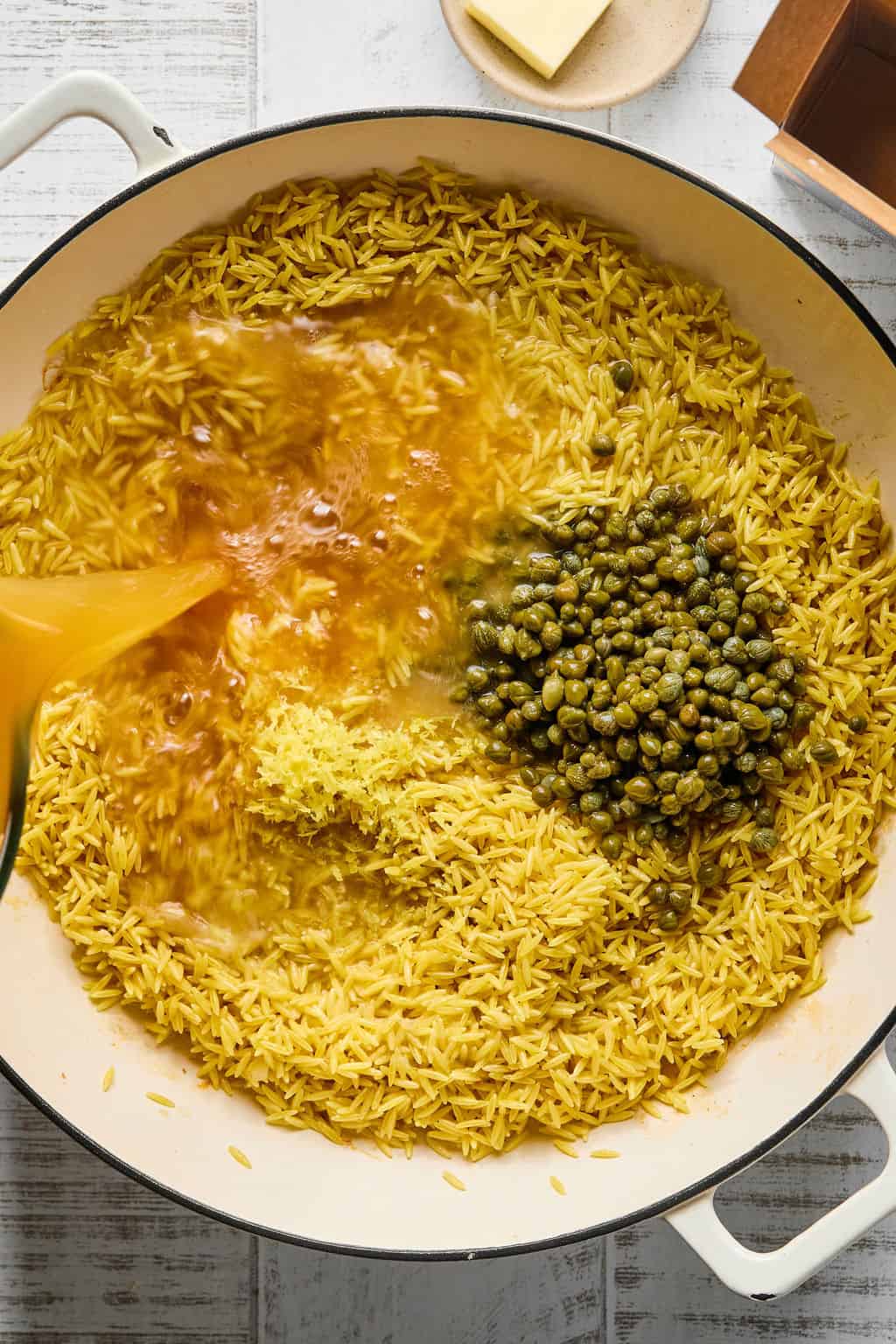 Adding capers and the caper juice from the jar, lemon juice, lemon zest, wine and broth to the orzo in pan