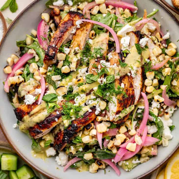 Grilled chicken salad with charred corn, pickled red onions, fresh greens, herbs, and crumbled cheese drizzled with a yellow dressing.