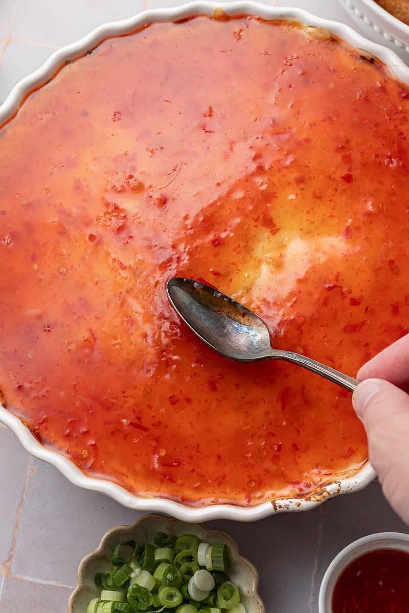 Spreading sweet chili sauce on top of Crab Rangoon Dip with a spoon.
