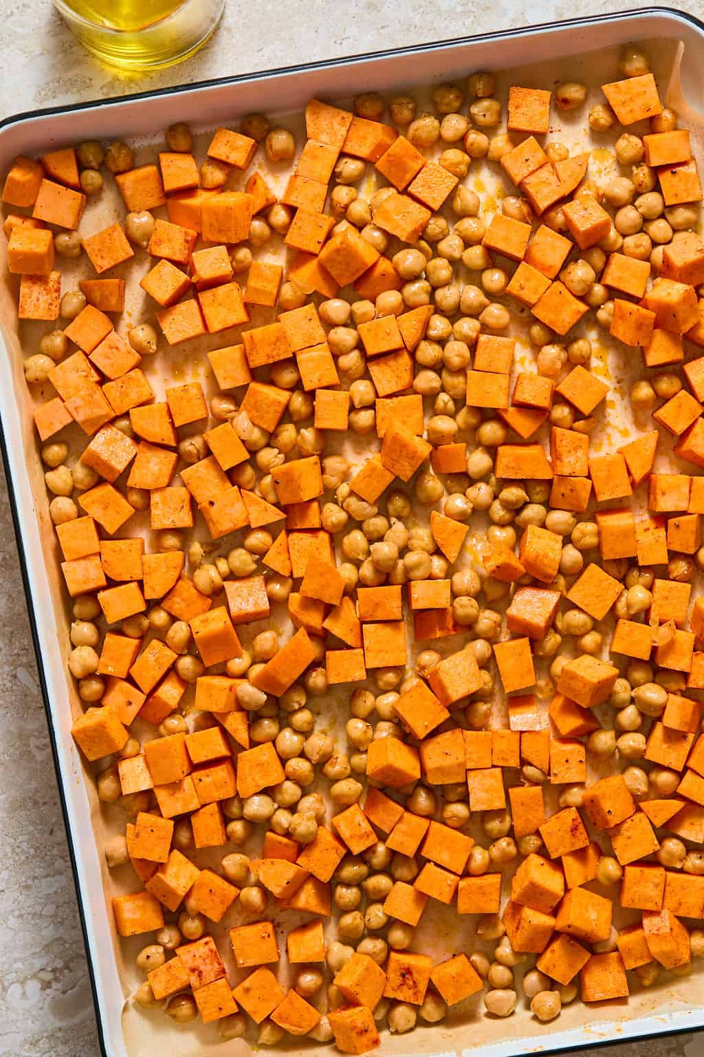 Overhead view of a sheet pan with diced sweet potato cubes and chickpeas seasoned with spices, ready to roast, with a small dish of oil in the corner.