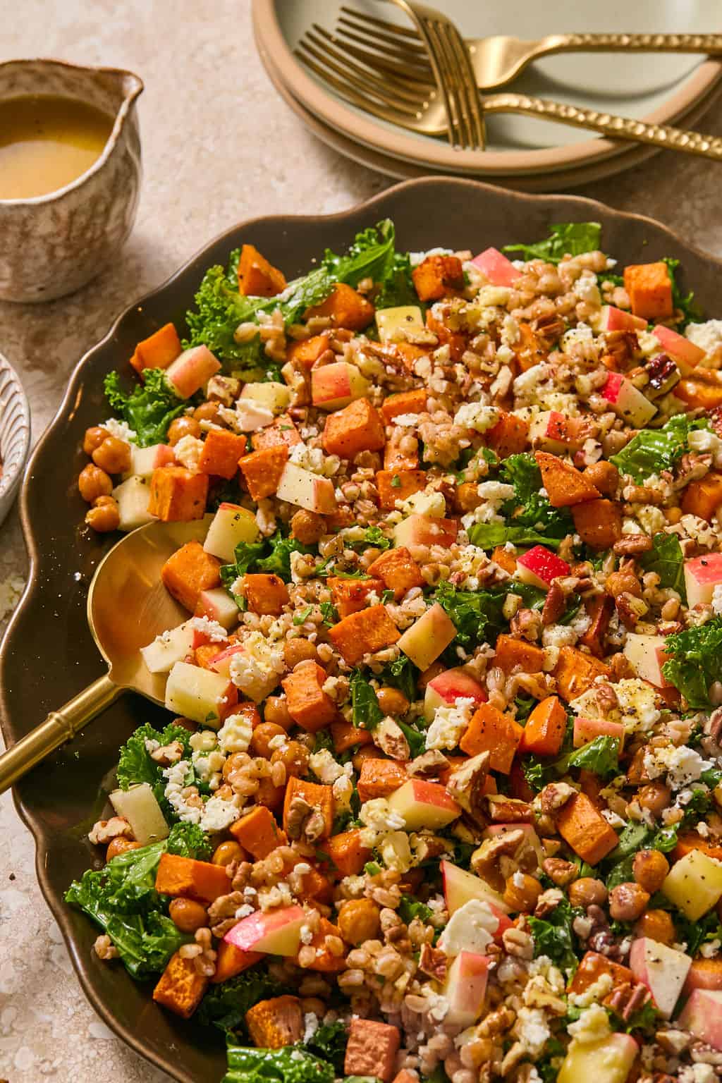 Overhead view of a kale salad with roasted sweet potato, chickpeas, farro, diced apple, feta, and pecans on a dark scalloped platter with a gold serving spoon, dressing and gold forks nearby.