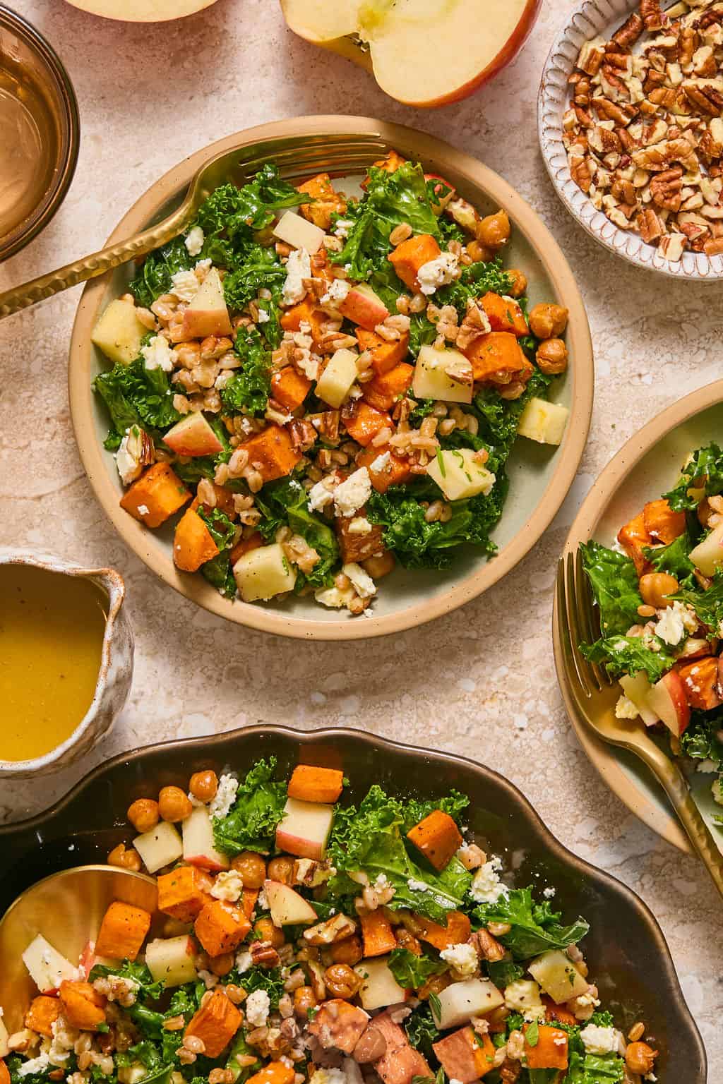 Two individual plates of kale salad with sweet potato, chickpeas, farro, apple, feta, and pecans, next to the serving platter, a small pitcher of dressing, a bowl of pecans, and apple halves.