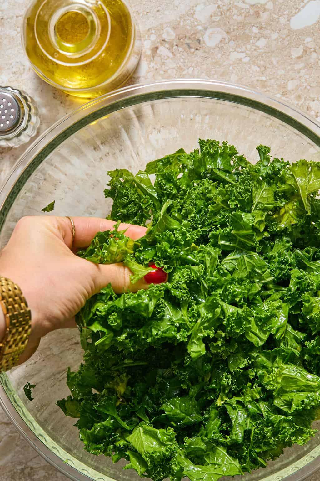 A hand massaging chopped kale in a large glass mixing bowl, with olive oil and a shaker of salt or pepper nearby on the counter.