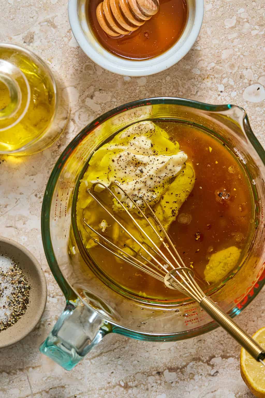 Overhead view of honey mustard vinaigrette being whisked in a glass measuring cup, with a small dish of honey, olive oil, cracked pepper, and a lemon half nearby.
