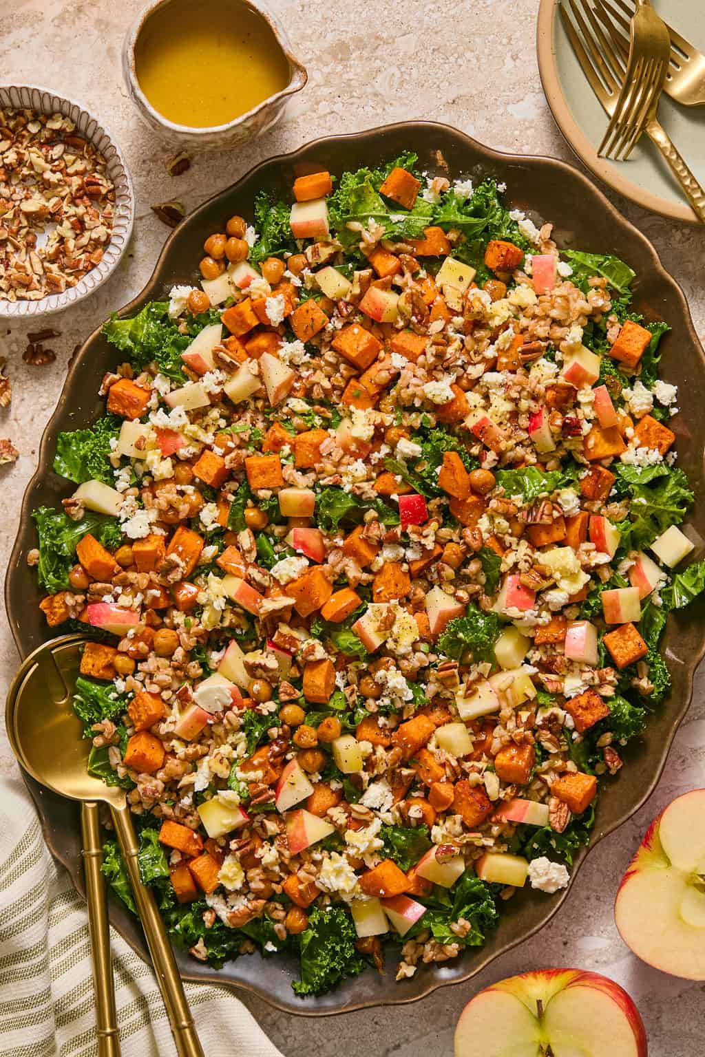 Overhead view of a fall farro salad on a dark scalloped platter, topped with roasted sweet potato cubes, farro, chickpeas, diced apple, crumbled feta, and chopped pecans, served with a side of yellow vinaigrette and a bowl of pecans, gold forks and a striped napkin nearby.